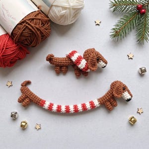 May include: Two brown crocheted dachshund figures, one small and one long, both wearing red and white striped scarves. The figures feature dark eyes and white muzzles. They are arranged with yarn skeins, small wooden stars, and silver jingle bells.