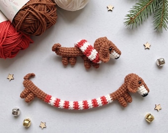 May include: Two brown crocheted dachshund figures, one small and one long, both wearing red and white striped scarves. The figures feature dark eyes and white muzzles. They are arranged with yarn skeins, small wooden stars, and silver jingle bells.