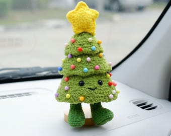 May include: A green crocheted Christmas tree figurine with a yellow star on top. The tree features small colorful pom-poms as ornaments, black bead eyes, a stitched smile, and two green crocheted legs standing on a small circular wooden base.