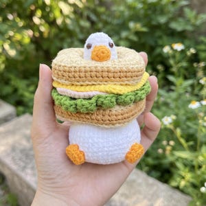 Può includere: Un giocattolo a forma di anatra fatto a mano all'uncinetto con un design a sandwich. La testa dell'anatra spunta da un panino marrone, giallo e verde. L'anatra ha un corpo bianco, zampe arancioni e un becco giallo. Gli strati del panino includono un panino marrone, ripieno giallo e lattuga verde.