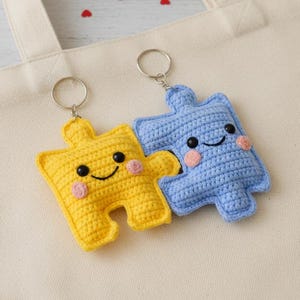 May include: Two crocheted puzzle piece keychains, one yellow and one blue, with smiling faces and pink cheeks, rest on a cream-colored tote bag. A red rose and a small card with text are in the background.