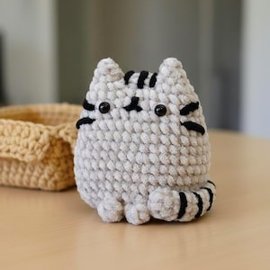 May include: A handmade, gray crocheted cat with black stripes and facial details. The plush cat has a rounded body, small ears, and a striped tail. A beige crocheted basket is in the background.