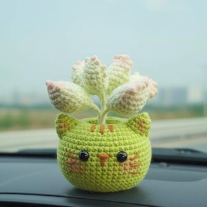 May include: A handmade crochet cat planter in light green, holding a succulent plant with light green and pink leaves. The cat planter has orange stripes and a cute face. The planter is sitting on a car dashboard.