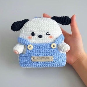 Può includere: Custodia a forma di cane all'uncinetto fatta a mano, con testa bianca, orecchie nere e salopette blu. La custodia ha un occhio che strizza e una piccola etichetta che dice "HANDMADE limited edition". La custodia è tenuta in una mano.