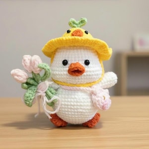 May include: A crocheted duckling toy, wearing a yellow hat with a green leaf, a pink scarf, and a small pink bag. It holds a bouquet of pink tulips. The duckling has black eyes, an orange beak and orange feet.