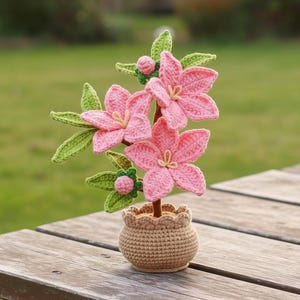 Op de afbeelding: Een handgemaakt gehaakt bloemstuk met roze bloemen, groene bladeren en knoppen, in een kleine, bruin gehaakte pot. De bloemen hebben gele centra en zijn gerangschikt op een bruine stengel.