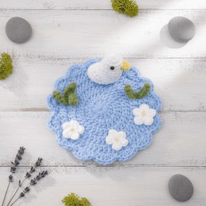 Peut inclure: Un dessous de verre fait à la main en crochet bleu clair avec un canard blanc, des feuilles vertes et des fleurs blanches. Le dessous de verre rond a un bord festonné. Le canard a un bec jaune et des yeux noirs. Des éléments décoratifs entourent le dessous de verre.