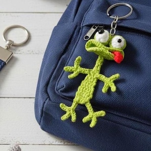 May include: A handmade green crochet frog keychain with white and black eyes and a red tongue. The frog is attached to a silver keyring and hangs on a navy blue backpack. A small, gray crocheted ball is also visible.