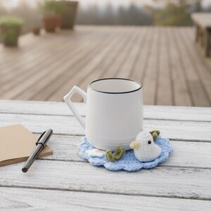 May include: A white ceramic mug with a black rim sits on a light blue crocheted coaster with a white duck design. A black pen and a small notepad are on a white wooden table. The background is a blurred outdoor scene.