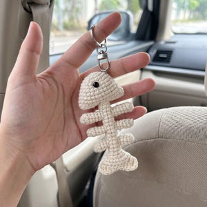May include: A cream-colored crocheted fish skeleton keychain featuring a single black bead eye. The fish bone structure is attached to a silver-toned metal split ring and a lobster clasp connector, creating a dangling accessory for keys or bags.