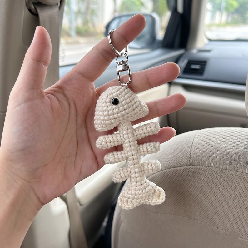 May include: A cream-colored crocheted fish skeleton keychain featuring a single black bead eye. The fish bone structure is attached to a silver-toned metal split ring and a lobster clasp connector, creating a dangling accessory for keys or bags.