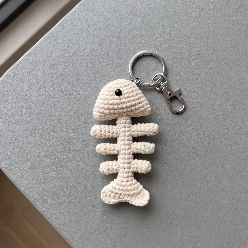 May include: A cream-colored crochet fish skeleton keychain featuring a single black bead for an eye and a silver-toned metal split ring with a lobster clasp attachment. The yarn structure is visible throughout the entire fish bone design.