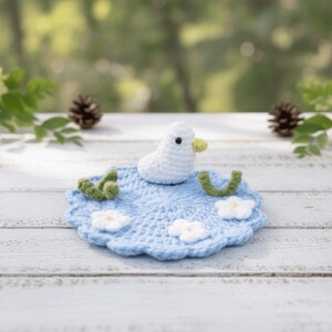 May include: A crocheted white duck with a yellow beak sits on a light blue crocheted mat with white flowers and green accents. The mat is round with a scalloped edge. The background is a blurred view of greenery.