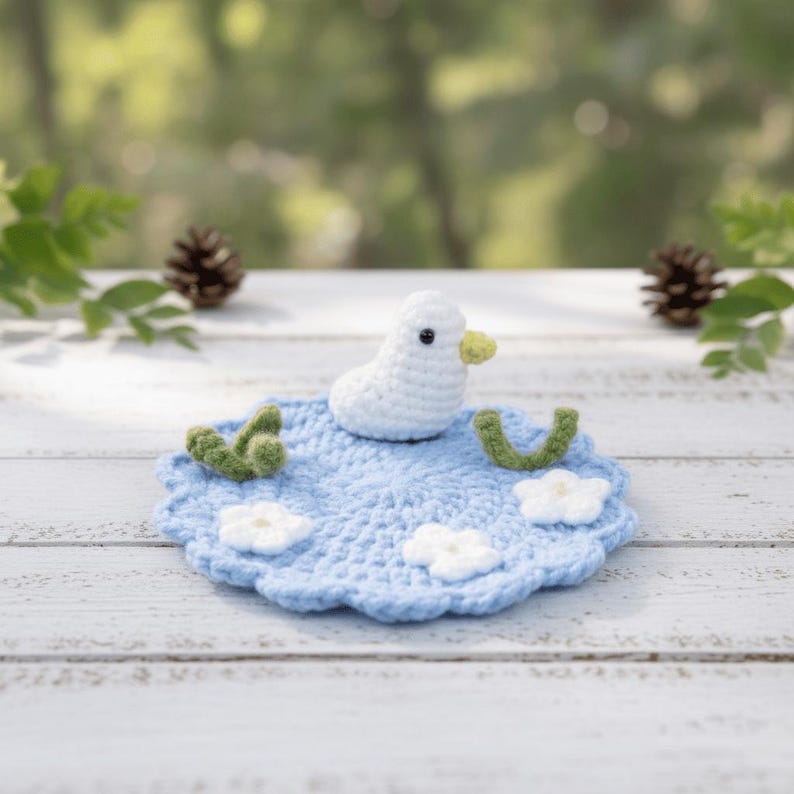 May include: A crocheted white duck with a yellow beak sits on a light blue crocheted mat with white flowers and green accents. The mat is round with a scalloped edge. The background is a blurred view of greenery.