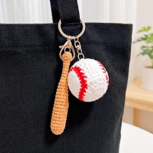 Könnte beinhalten: Ein Schlüsselanhänger mit einem kleinen, beigefarbenen gehäkelten Baseballschläger und einem weißen gehäkelten Baseball mit roten Nähten. Die beiden Anhänger sind an einem silberfarbenen Ring und Karabiner an einer schwarzen Stofftasche befestigt.