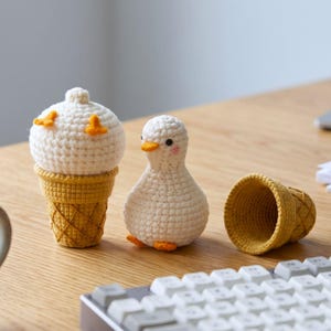 May include: A white crocheted duck figurine standing next to a white crocheted ice cream scoop in a yellow waffle cone, with an empty yellow crocheted waffle cone lying on its side nearby, all crafted with visible yarn stitches and textures.