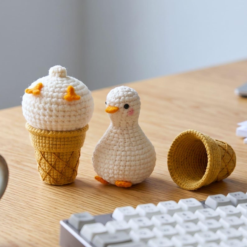 May include: A white crocheted duck figurine standing next to a white crocheted ice cream scoop in a yellow waffle cone, with an empty yellow crocheted waffle cone lying on its side nearby, all crafted with visible yarn stitches and textures.