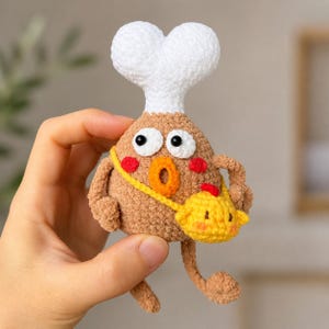 May include: A handmade, brown crochet chicken leg character with a white chef's hat, large eyes, and a yellow purse. The character has red cheeks and a small yellow purse with a red accent. The design is whimsical and playful.