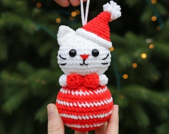 May include: A handmade crochet Christmas ornament shaped like a cat wearing a Santa hat and bow tie. The cat is white with black eyes and whiskers, and the hat and bow tie are red. The ornament's body is a red and white striped ball.