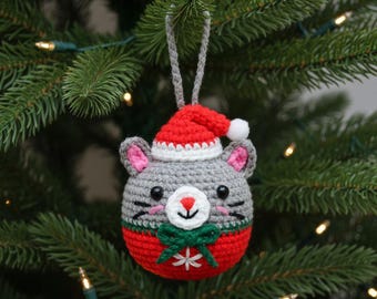 May include: A handmade crochet Christmas ornament shaped like a gray mouse wearing a red Santa hat with a white pom-pom. The mouse has pink ears, a red nose, and a green bow. A white snowflake is on the red bottom half.