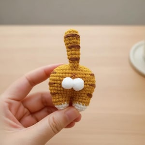 Op de afbeelding: Een handgemaakte gehaakte kattenkont in tinten geel en bruin. De achterkant van de kat heeft twee pluizige witte ballen en een gestreepte staart. De onderkant van de kattenpoten is wit.