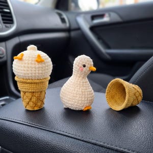 May include: A crochet duck toy with a round cream-colored body, small orange feet, and pink cheeks, positioned next to a cream-colored crochet scoop inside a golden-brown waffle cone and an empty matching waffle cone with a textured grid pattern.