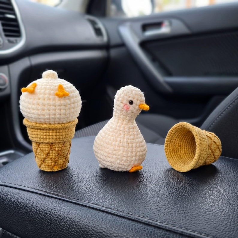 May include: A crochet duck toy with a round cream-colored body, small orange feet, and pink cheeks, positioned next to a cream-colored crochet scoop inside a golden-brown waffle cone and an empty matching waffle cone with a textured grid pattern.
