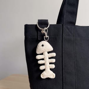May include: A cream-colored crocheted fish skeleton keychain attached to a black canvas tote bag with a silver-toned metal lobster clasp and a circular ring. The fish bone design features a single small black bead for an eye on the head.