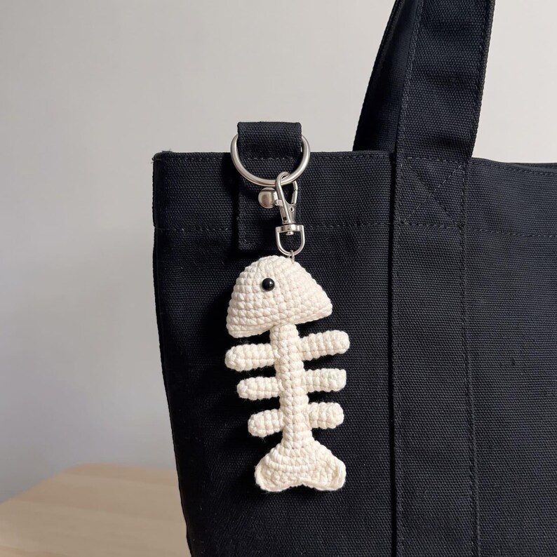 May include: A cream-colored crocheted fish skeleton keychain attached to a black canvas tote bag with a silver-toned metal lobster clasp and a circular ring. The fish bone design features a single small black bead for an eye on the head.