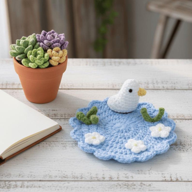 May include: A terracotta pot holds a cluster of crocheted succulents in shades of green, purple, and yellow. A crocheted blue coaster with a white duck, green leaves, and white flowers sits nearby. A notebook is open on the left.