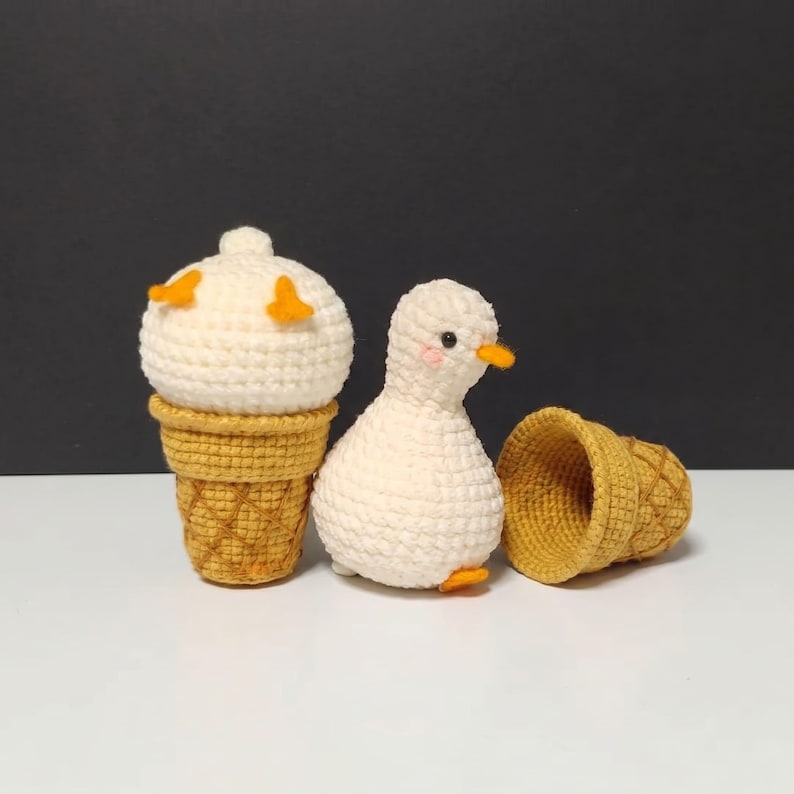 May include: A crocheted white duck figurine with orange feet and beak, positioned next to a crocheted ice cream cone with a white scoop and a yellow waffle cone, and a separate empty yellow crocheted waffle cone lying on its side.