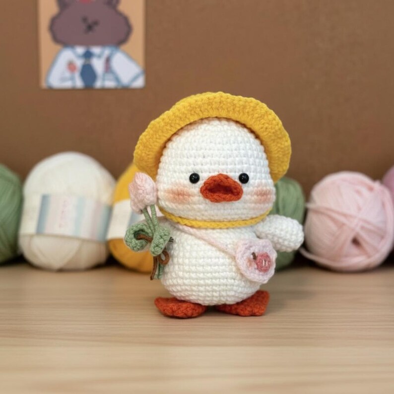 May include: A crocheted duck toy, white with a yellow hat and a pink purse, holding a bouquet of pink flowers. It has orange feet and a yellow neckband. Balls of yarn in various colours are in the background.