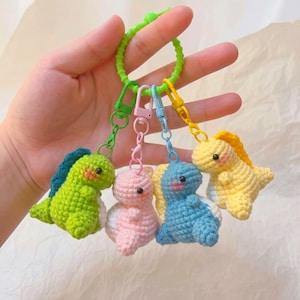 May include: A set of four small, crocheted dinosaur keychains in green, pink, blue, and yellow, each attached to a matching colored plastic lobster clasp. The four clasps are connected to a single, larger, bright green circular plastic ring.