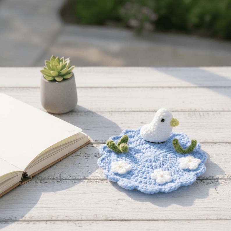 May include: A handmade crochet coaster featuring a white duck on a blue water-themed design. The coaster has white flower accents and green leaf details. A small succulent in a gray pot and an open book are also visible.