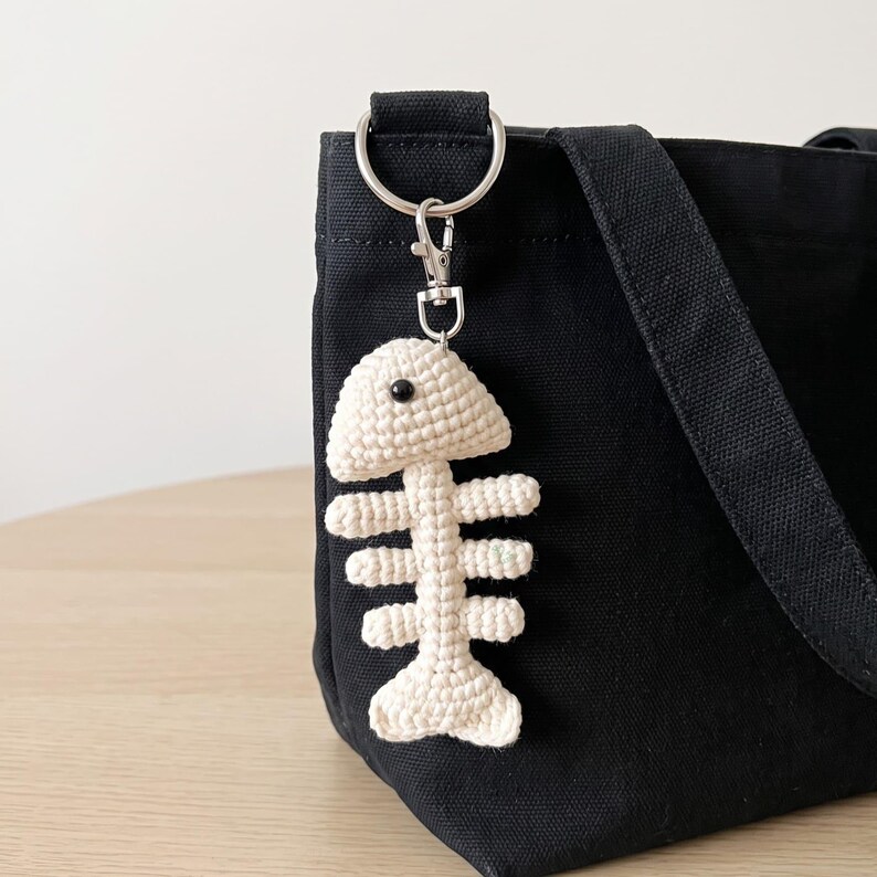 May include: A cream-colored crocheted fish skeleton keychain attached to a silver-toned metal lobster clasp and ring, hanging from the side of a black canvas tote bag. The fish bone charm features a single small black bead for an eye.