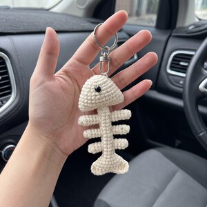 May include: A cream-colored crochet fish skeleton keychain featuring a small black bead for an eye. The fish bone design is crafted from yarn and attached to a silver-toned metal split ring and a lobster clasp for secure attachment to keys.