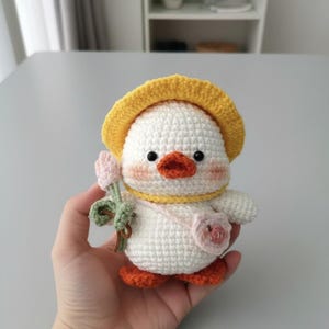 May include: A crocheted duckling toy, white with orange feet and beak, wearing a yellow hat and holding a pink flower. It has a yellow collar and a small pink purse. The duckling has pink cheeks.