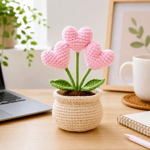May include: A handcrafted crochet flower arrangement with three pink heart-shaped flowers, green stems and leaves, in a beige crochet pot. The decorative piece is placed on a wooden surface. The arrangement is a charming, whimsical design.