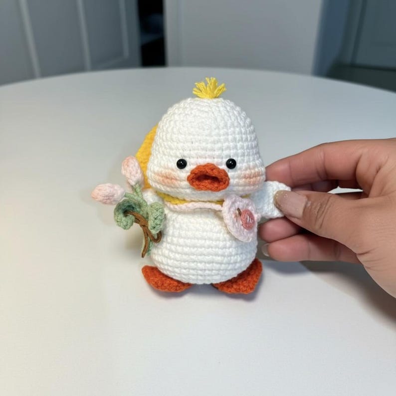May include: A crocheted duckling figurine with a white body, orange feet and beak, and black eyes. The duckling has a yellow tuft of hair and a yellow scarf. It holds a sprig of pink flowers and green leaves.