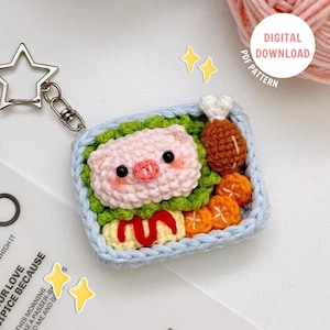 Pode incluir: Um porta-chaves de croché com um design de caixa bento retangular e rebordo azul claro. No interior, são visíveis uma cara de porco cor-de-rosa, alface verde, uma coxa de frango castanha, rodelas de cenoura laranja e tamagoyaki amarelo com molho vermelho. Inclui um fecho em forma de estrela.