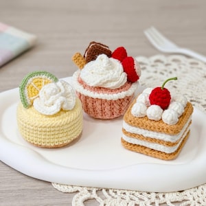 May include: Three crocheted dessert replicas: a round yellow cake topped with a lime slice and white cream, a pink cupcake with white frosting and red berries, and a square tan cake layered with white cream and topped with a red cherry.