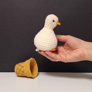 May include: A small, white, crocheted duck figurine with a round body, black eyes, a small orange beak, and pink cheeks, positioned above a separate, hollow, yellow, crocheted ice cream cone shape with a textured, waffle-like pattern.