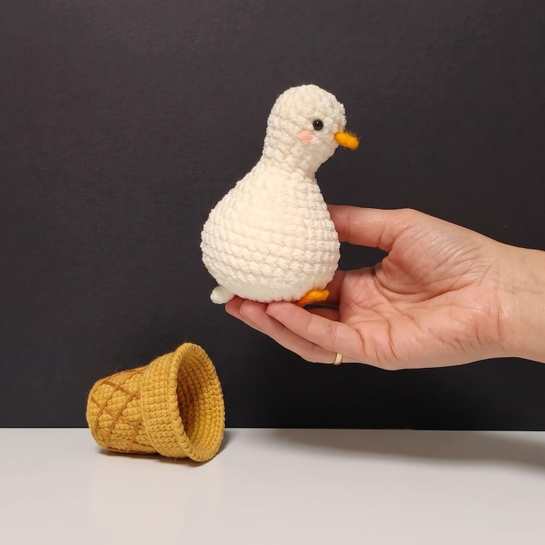 May include: A small, white, crocheted duck figurine with a round body, black eyes, a small orange beak, and pink cheeks, positioned above a separate, hollow, yellow, crocheted ice cream cone shape with a textured, waffle-like pattern.