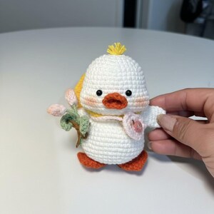 May include: A handmade white crochet duck with orange feet and beak, black eyes, and a yellow tuft of hair. The duck holds a sprig of pink flowers and a pink heart. It has a yellow scarf and blush-coloured cheeks.