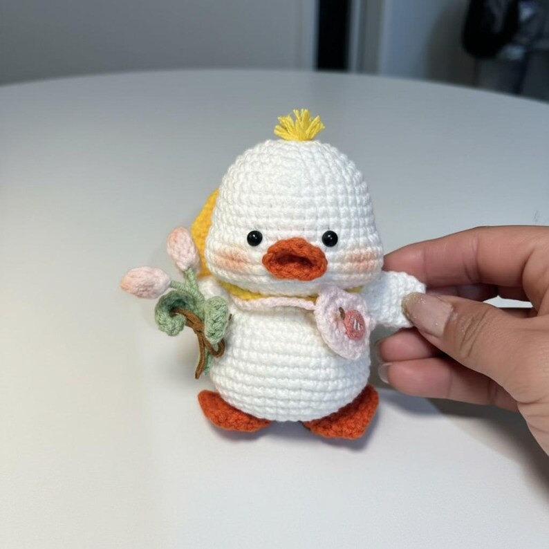 May include: A handmade white crochet duck with orange feet and beak, black eyes, and a yellow tuft of hair. The duck holds a sprig of pink flowers and a pink heart. It has a yellow scarf and blush-coloured cheeks.