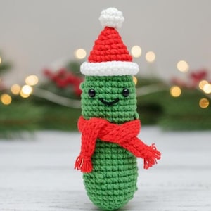 May include: A handmade, crocheted Christmas pickle ornament. The green pickle wears a red Santa hat with a white trim and a red scarf. The pickle has a smiling face with black eyes and a black mouth. The background is blurred with Christmas lights.
