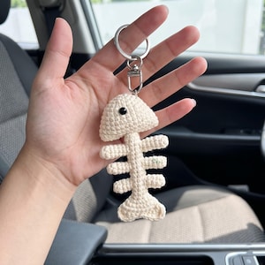 May include: A cream-colored crochet fish skeleton keychain featuring a small black bead eye. The fish bone design is attached to a silver-toned metal split ring and a lobster clasp, creating a textured, handmade accessory for keys or bags.