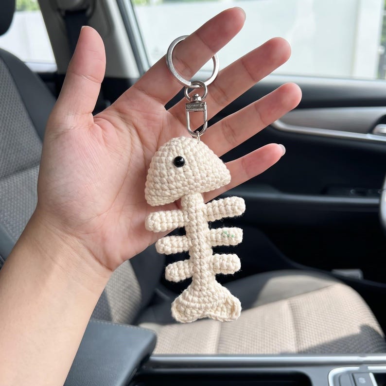 May include: A cream-colored crochet fish skeleton keychain featuring a small black bead eye. The fish bone design is attached to a silver-toned metal split ring and a lobster clasp, creating a textured, handmade accessory for keys or bags.