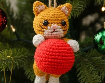 May include: A handmade crochet cat Christmas ornament. The cat is orange and white, holding a red ball. The ornament is hanging from a natural twine string. The background includes a Christmas tree with ornaments.