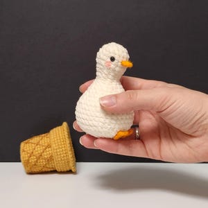 May include: A small, cream-colored crocheted duck toy with a bright orange beak and tiny pink blush marks on its cheeks. Beside it sits a separate, textured, golden-yellow crocheted ice cream cone cup with a cross-hatched pattern on its exterior.